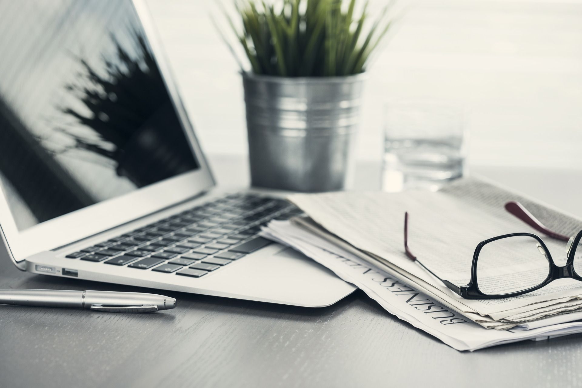 A laptop computer is sitting on a desk next to a newspaper and glasses.