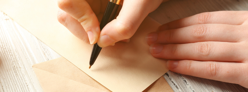 Hand holding a pen addressing an envelope and card sititng on a tble