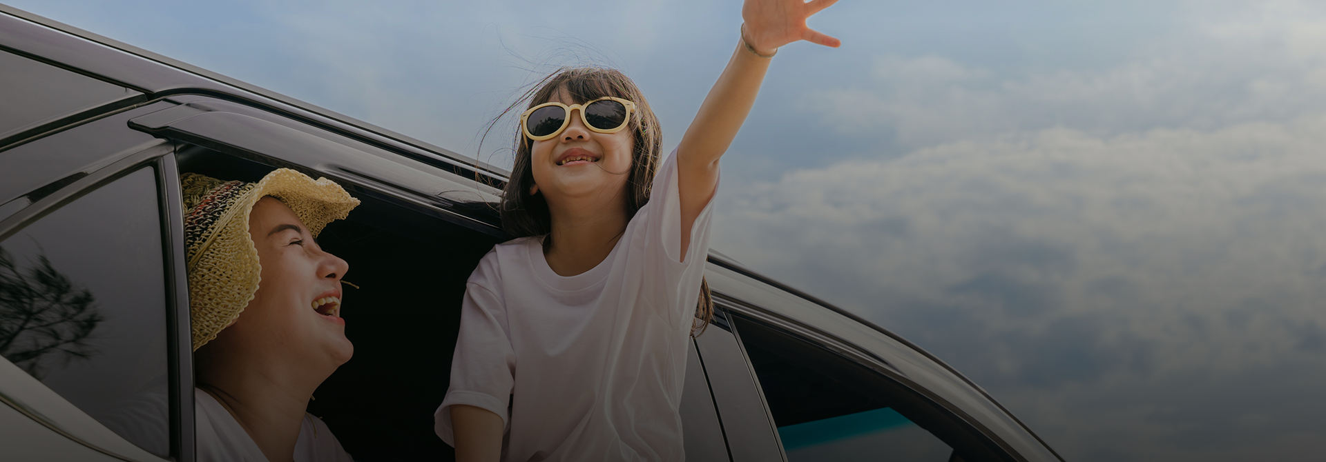 Two children are smiling and waving out the windows of a car, under a cloudy sky. One child wears sunglasses and raises their arm.