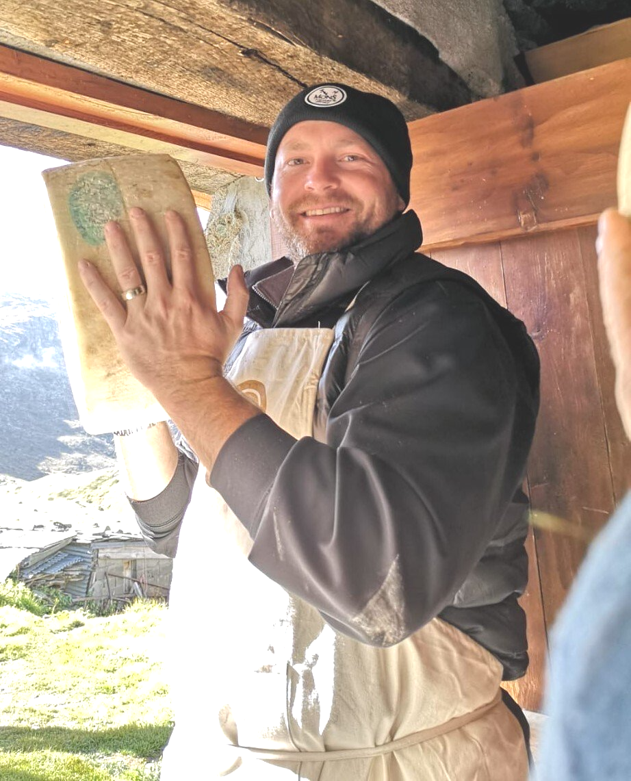 a man wearing a beanie and apron is holding a piece of cheese