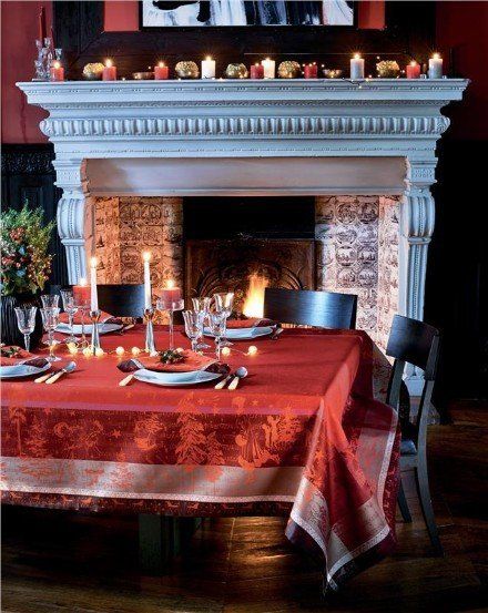 a table with a red table cloth and candles on it in front of a fireplace