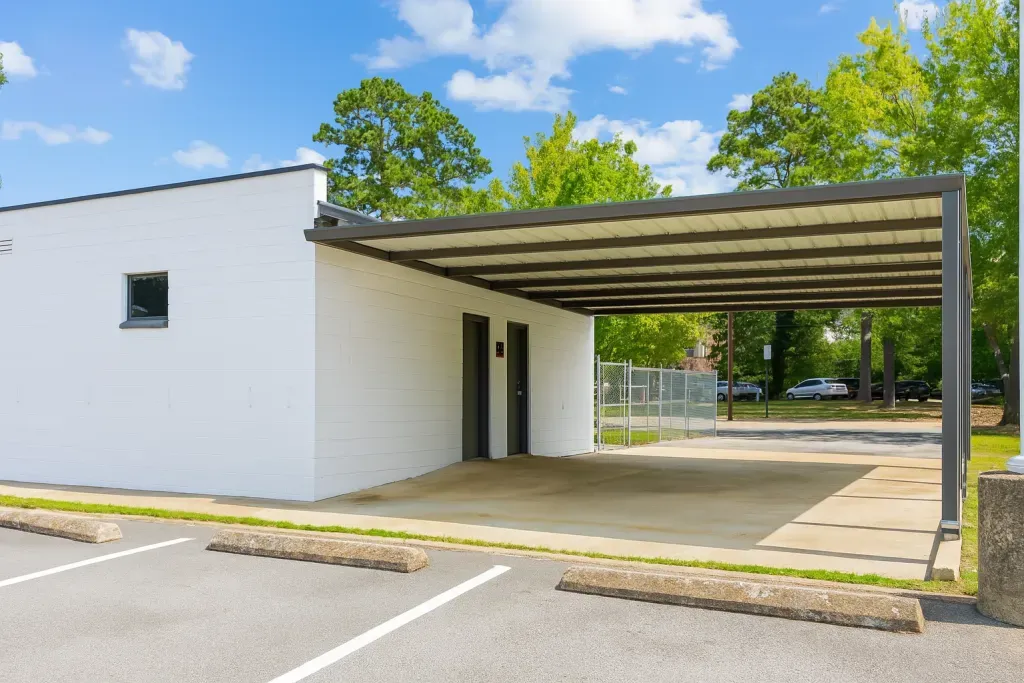 White building with gray carport, concrete parking area, and green trees under a blue sky.