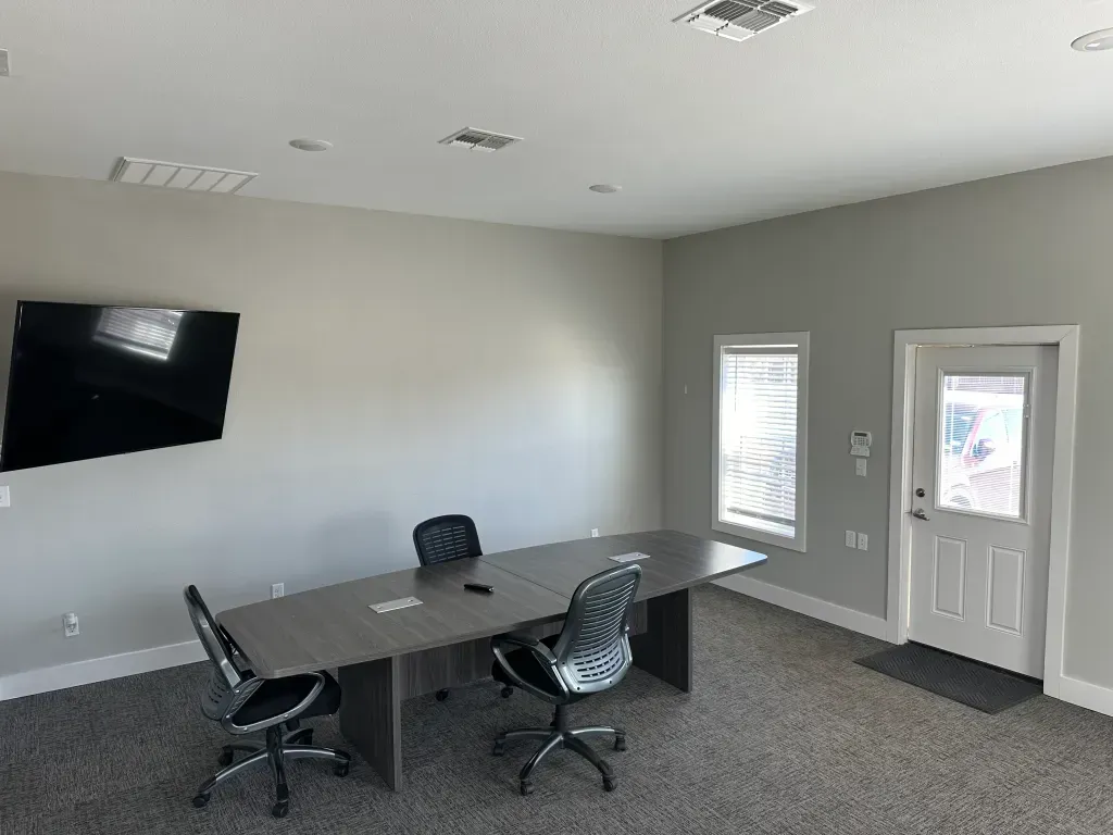Conference room with table, chairs, TV, windows, and door. Gray walls and carpet.