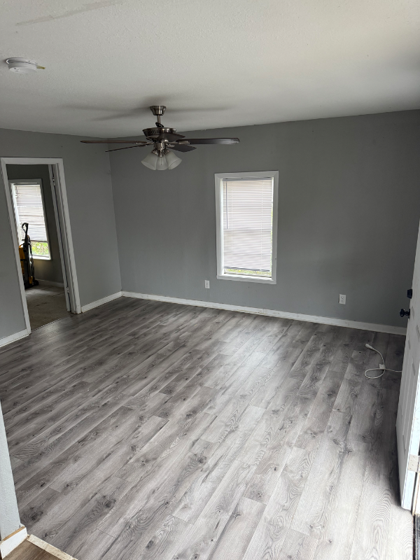 Gray-walled room with gray wood-look flooring. A window, door, and doorway are visible. A ceiling fan is mounted.