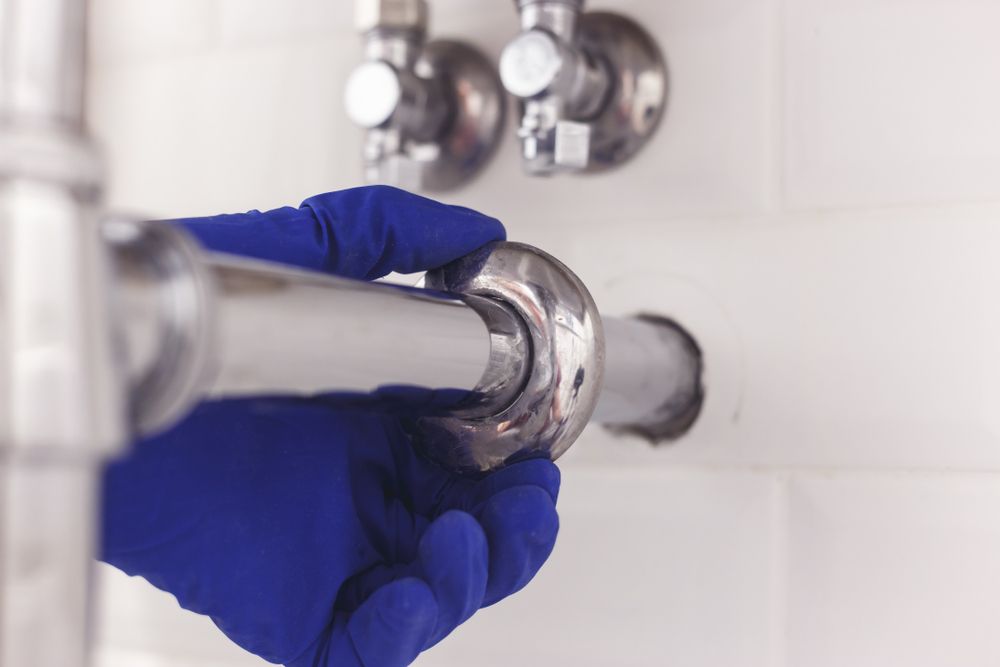 Gloved hand connecting or disconnecting shiny metal pipe under a sink. White tiled wall and chrome fixtures in background.