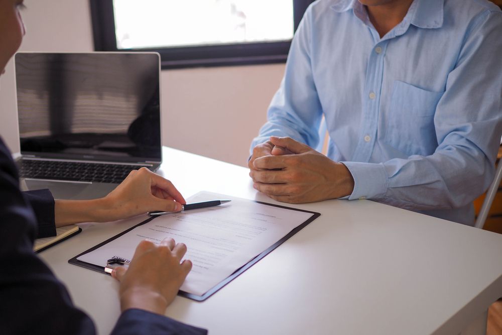 Person in blue shirt at a table with a clipboard, facing another person out of view, with a laptop in the background.
