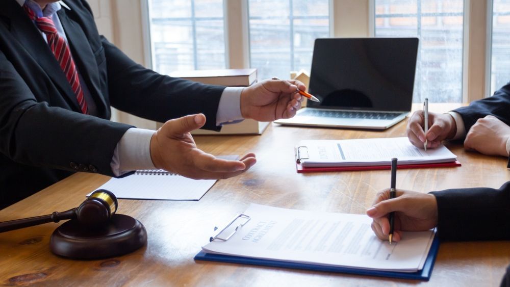 Lawyers in suits at a table with documents, a gavel, and a laptop. They are discussing and writing.