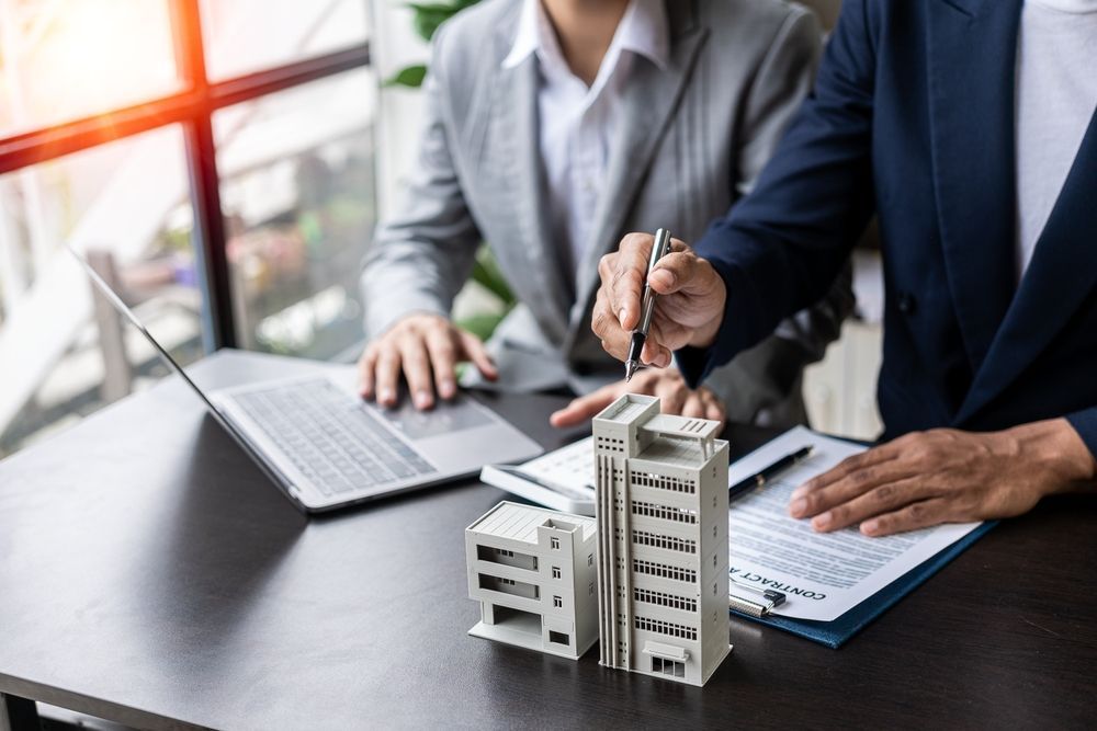 Two people in suits looking at a building model, pointing at papers on a desk with a laptop.