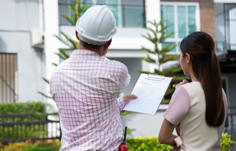 Home inspector in hard hat points at a house, woman looks on holding a pen, sunny day.