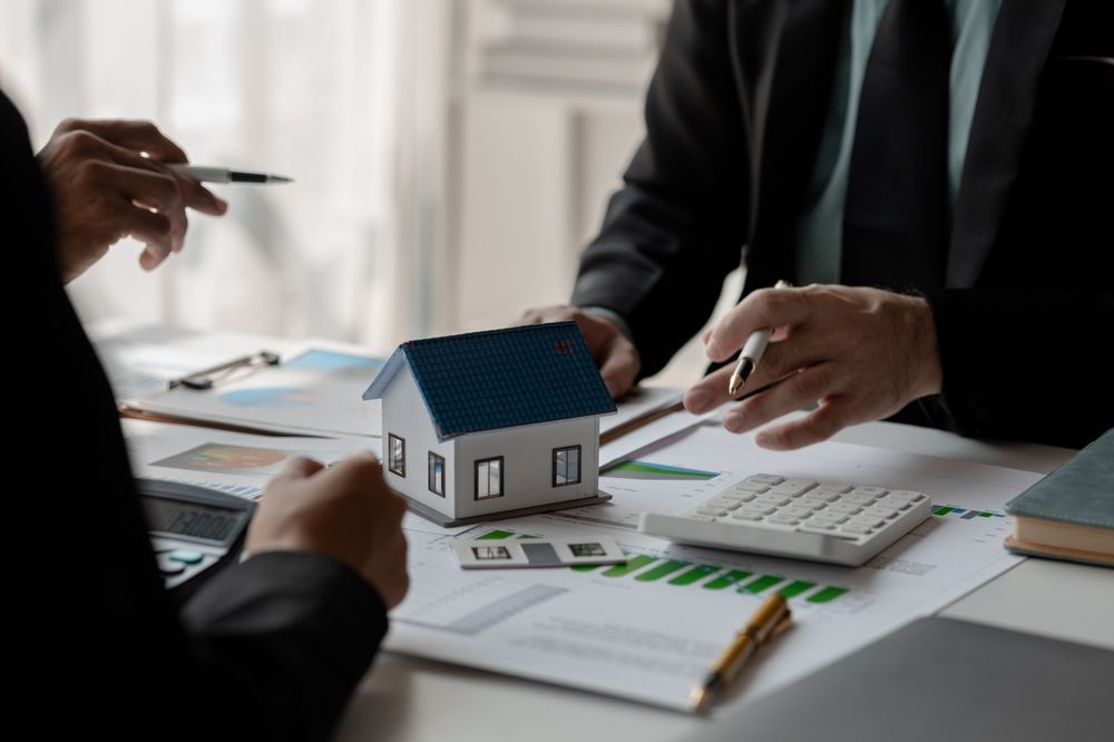 People in suits discussing documents around a miniature house model, calculator, and paperwork on a desk.