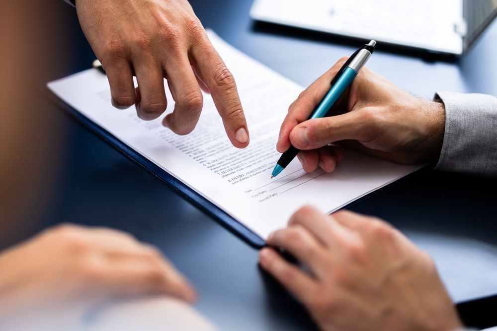 Hands signing a document. One hand points to a line while the other holds a pen.