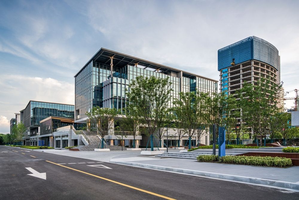 Modern glass and concrete office buildings with green trees and a paved street under a cloudy sky.