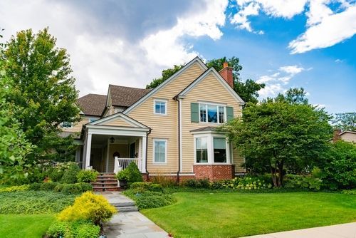Two-story beige house with a porch and green shutters, surrounded by a lawn and trees under a cloudy blue sky.