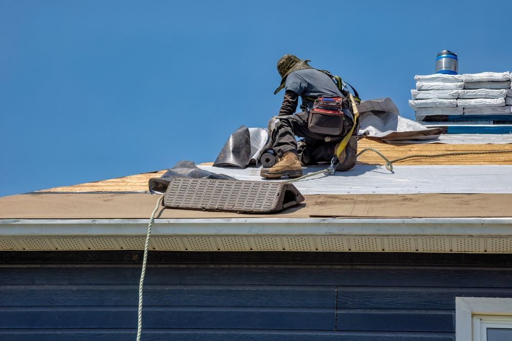 Roofer working on a roof, wearing a harness, against a blue sky.