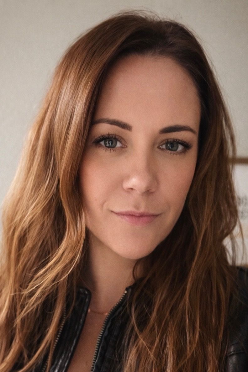 Football coach, arms crossed, on the field. Blue shirt, serious expression, players in background.