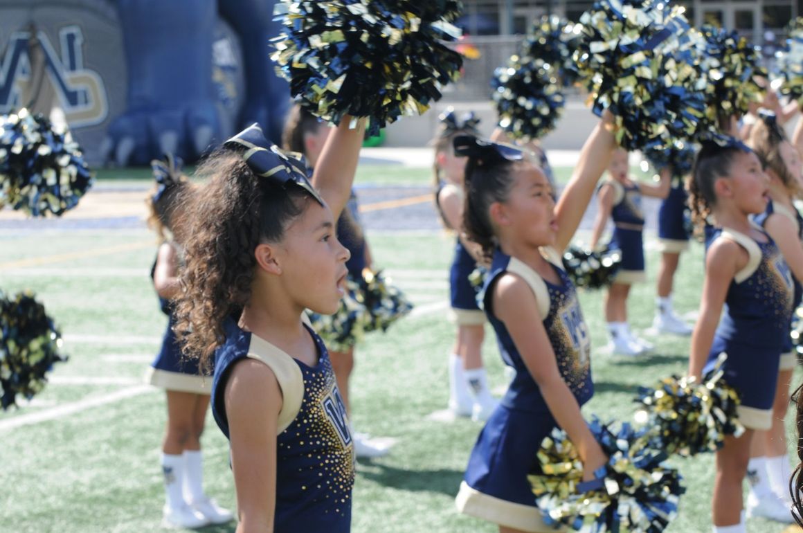 Football players in action on a grassy field; one runs with the ball, pursued by two opponents.