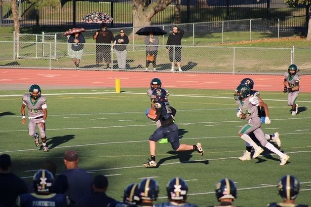 Flag football game at night, a player in red jersey runs with the ball as others pursue on a green field.