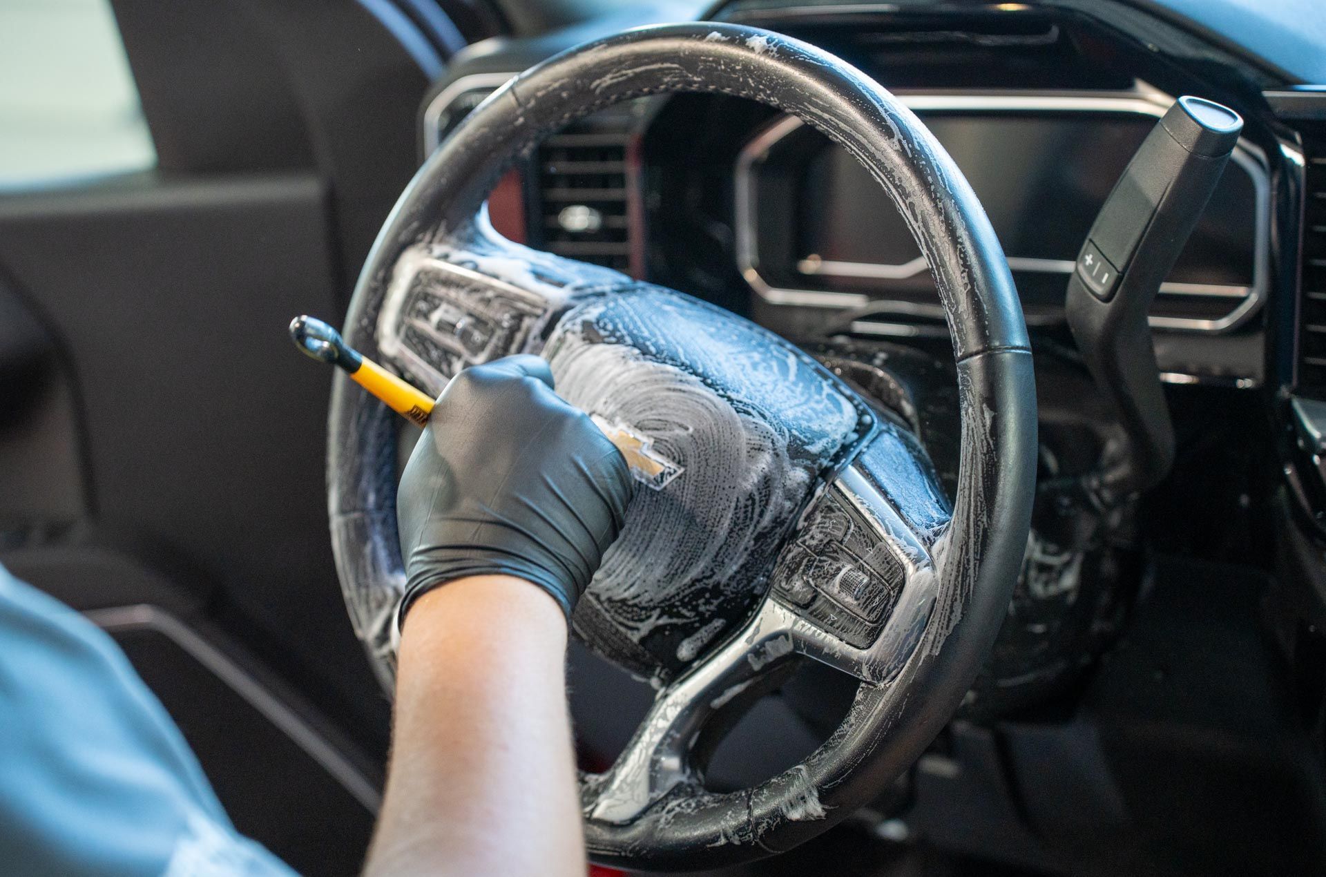 A hand wearing a black glove uses a small brush to apply cleaning foam to a car steering wheel.