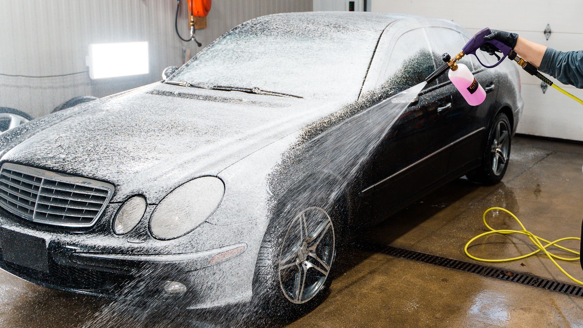 A person uses a foam cannon to apply thick white soap to the side of a black Mercedes-Benz in a garage.
