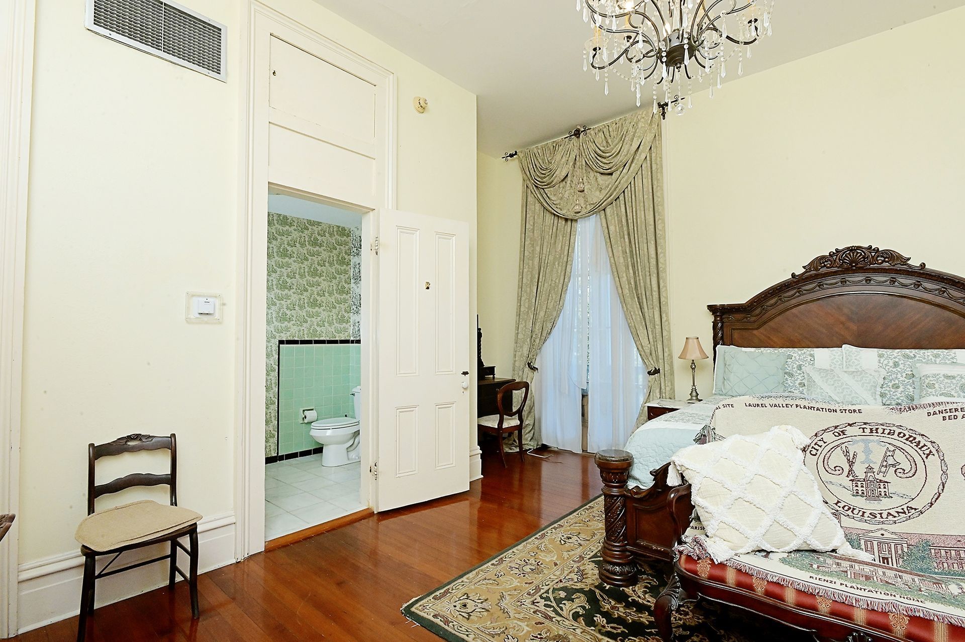 Bedroom with open bathroom door; ornate bed, chandelier, and wood floors.