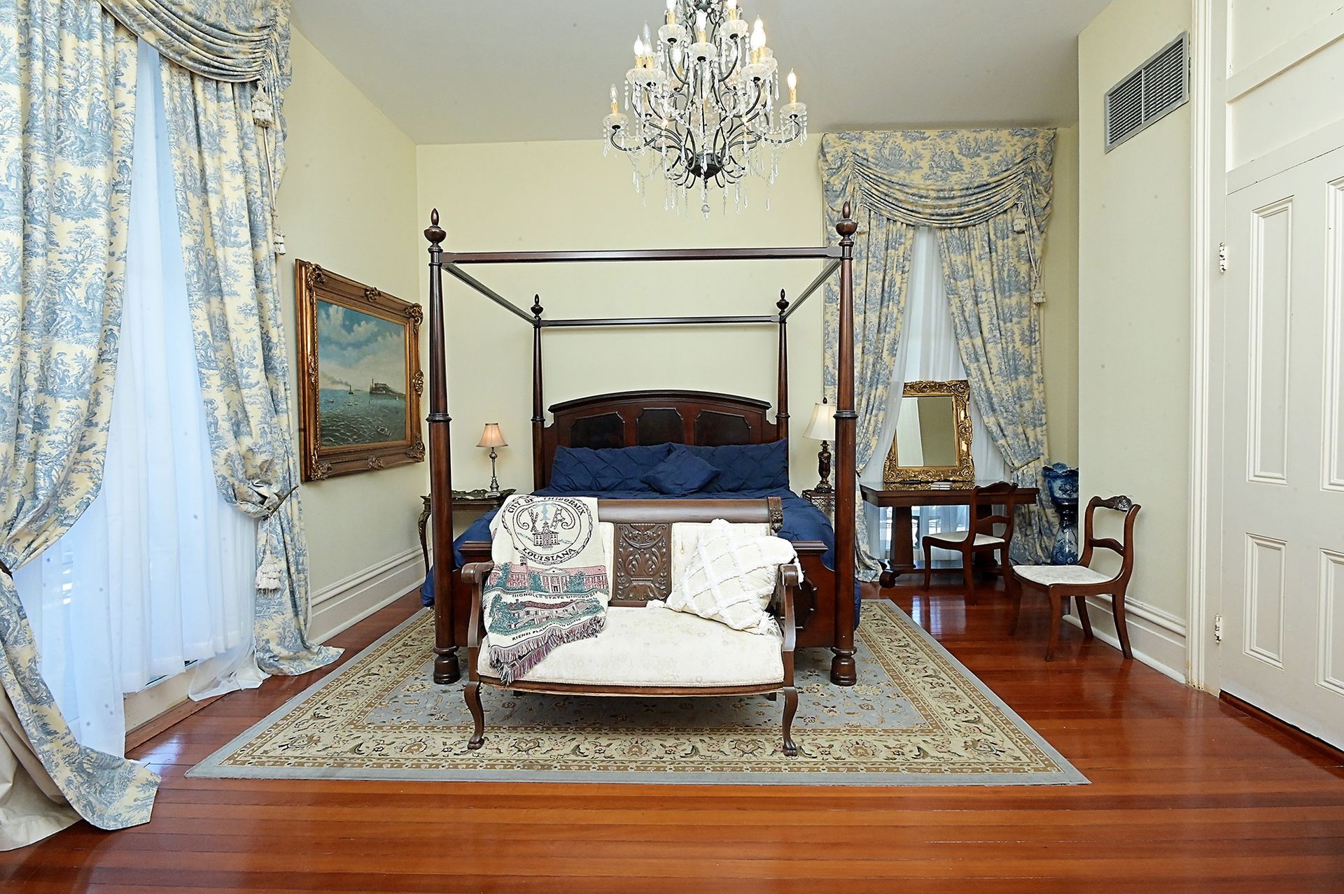 Elegant bedroom with four-poster bed, ornate curtains, antique furniture, and a chandelier.