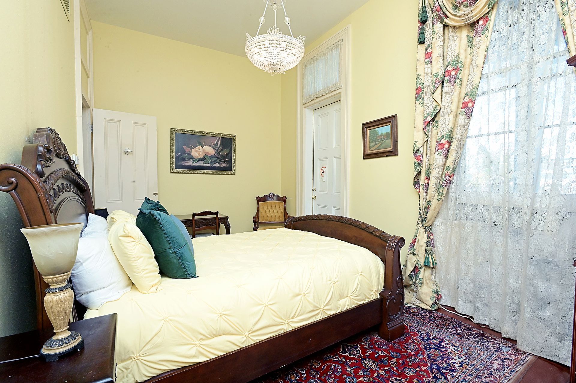 Bedroom with a dark wood bed, floral curtains, a rug, and a chandelier; yellow walls.