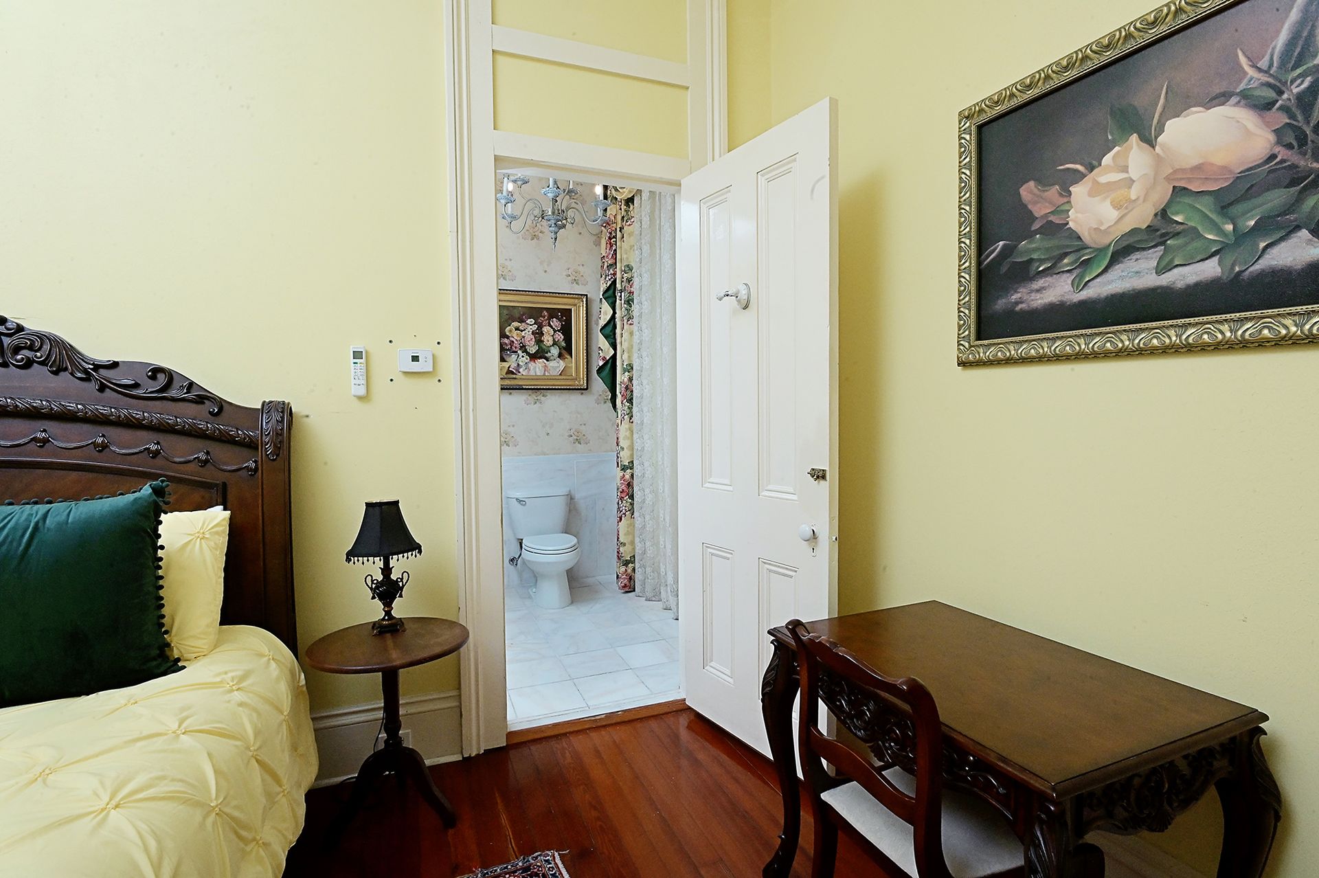 Bedroom with yellow walls, ornate bed, small table with lamp, desk, and open doorway to bathroom.