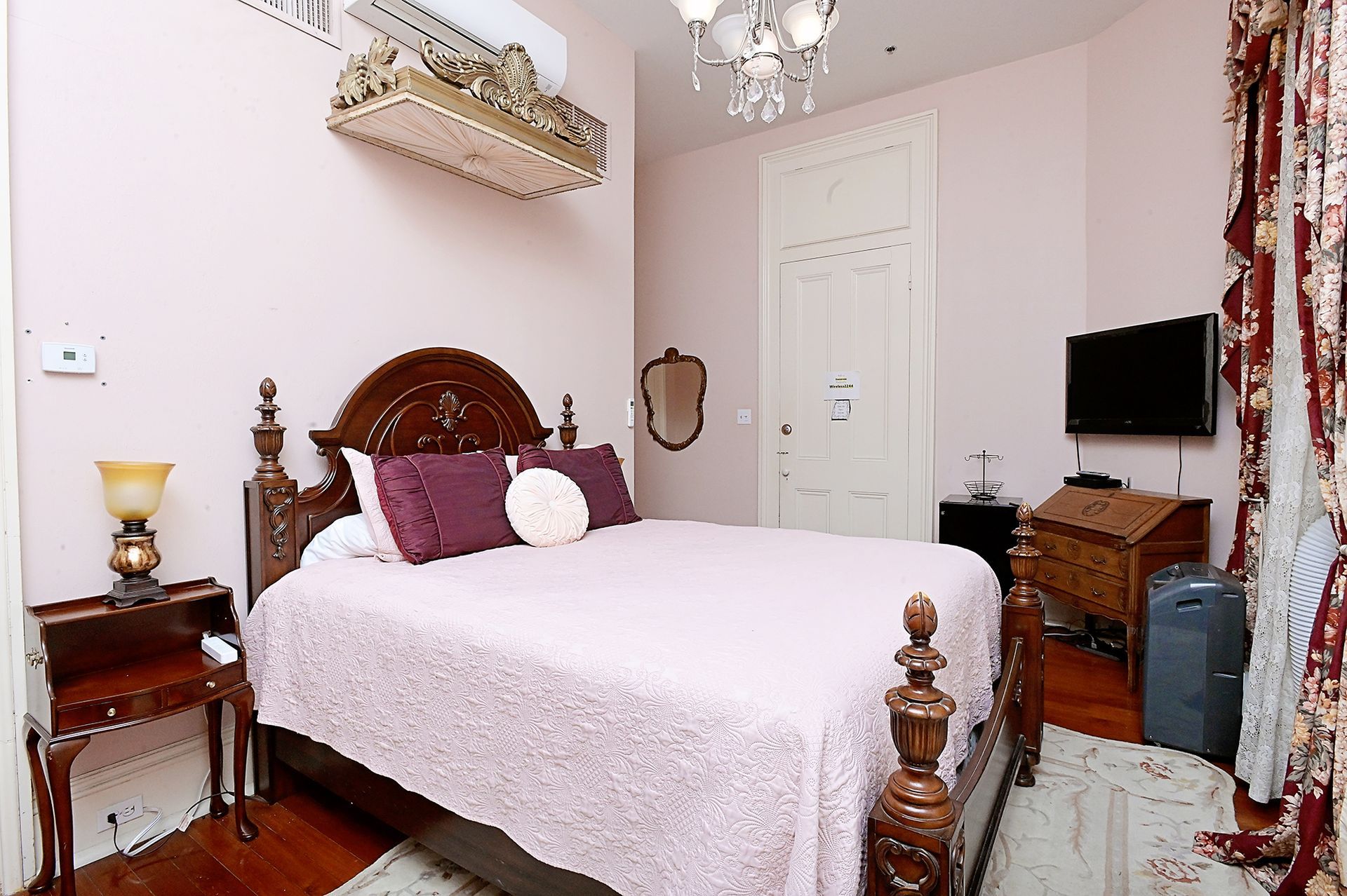 Bedroom with a queen bed, dark wood furniture, and pink walls. A small TV is mounted on the wall.