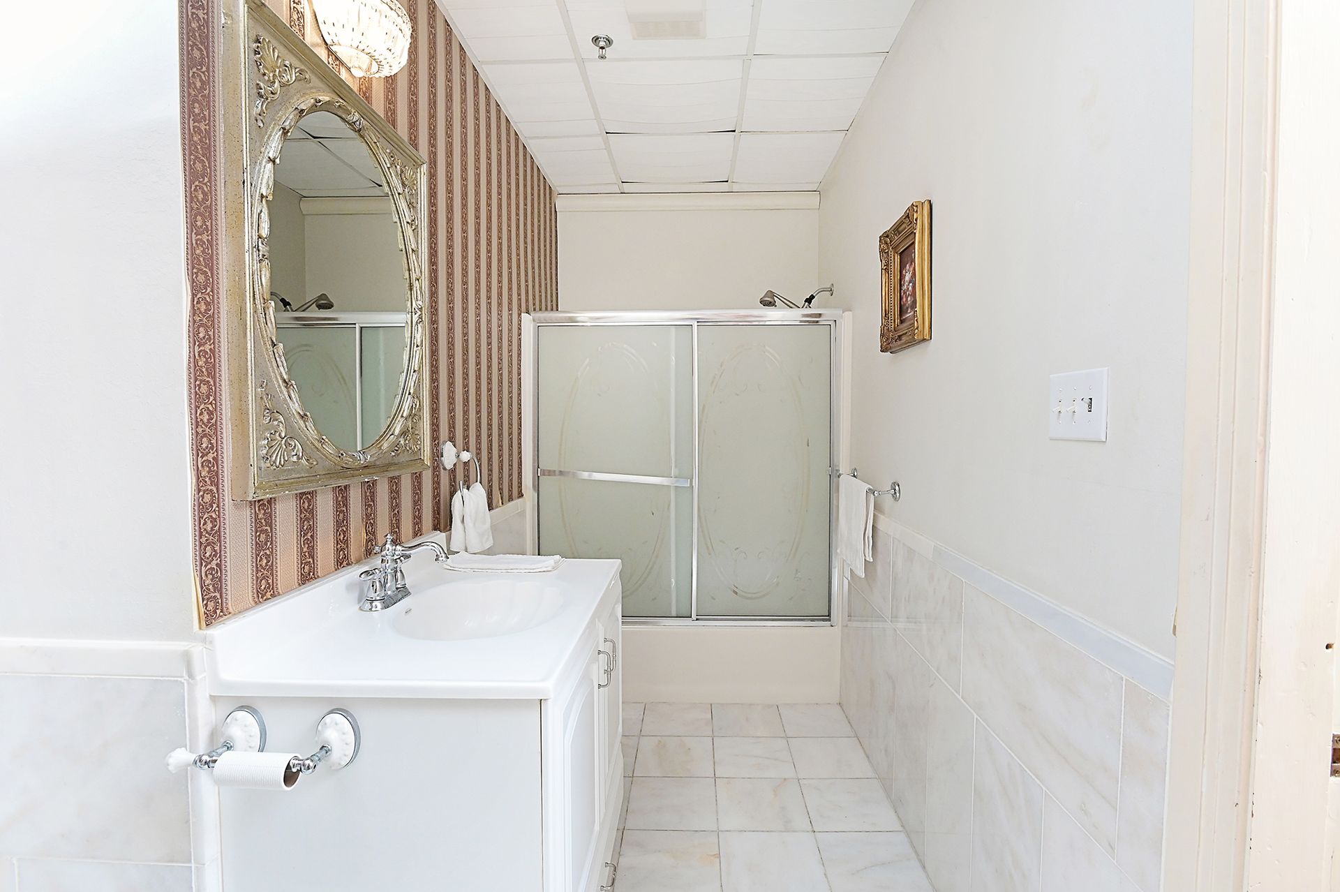 Bathroom with a vanity, shower, toilet paper holder, and ornate mirror. White and beige colors dominate.