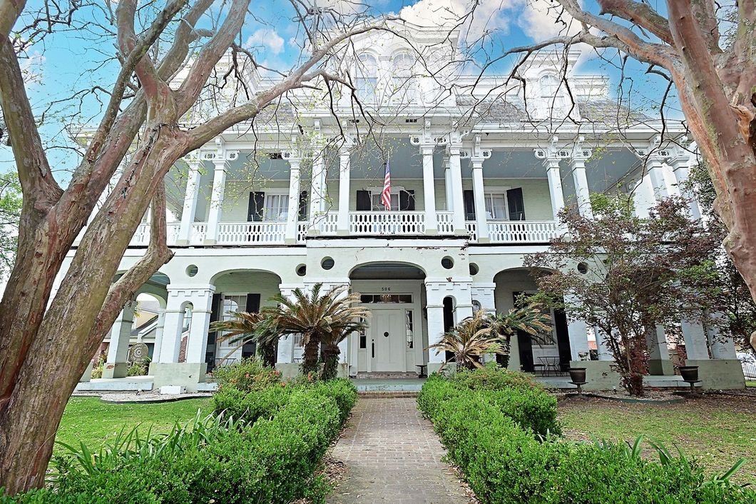 White Victorian house with two-story porch, fronted by a green lawn and a walkway lined with shrubs.