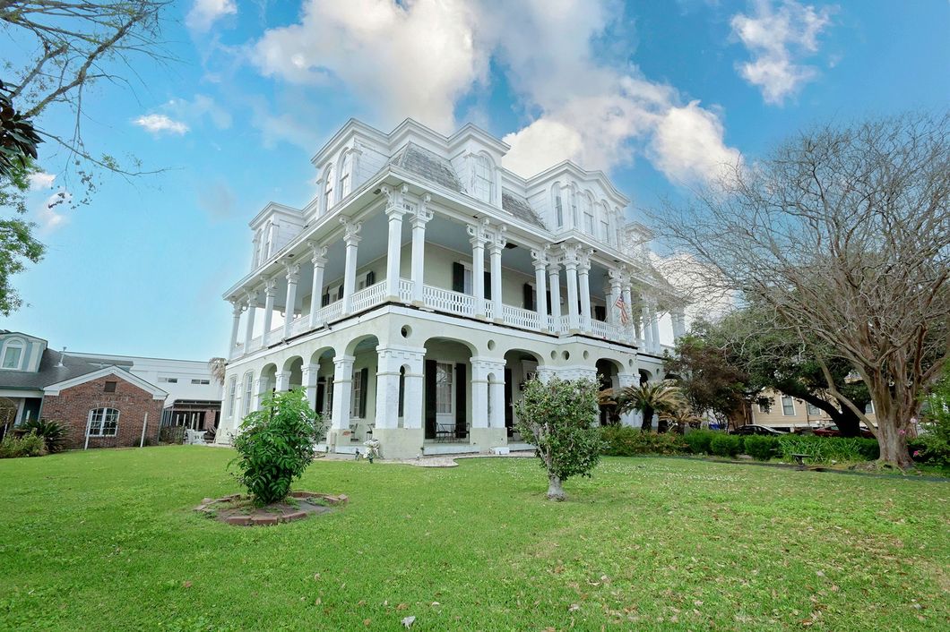 White Victorian mansion with columns, two-story porch, green lawn, and blue sky.