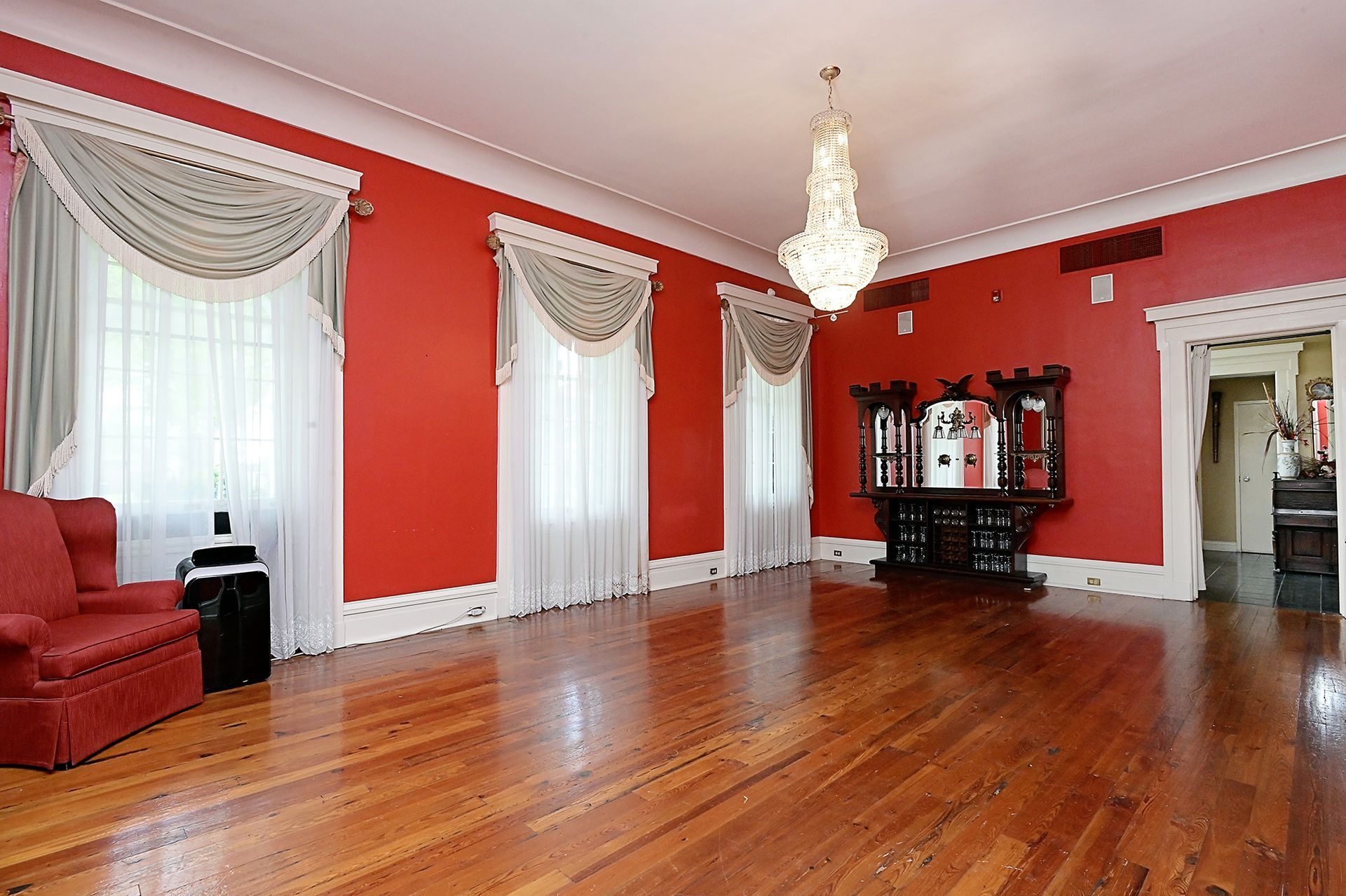Red-walled room with wood floor, white trim, and chandelier. Includes red furniture and a dark cabinet.
