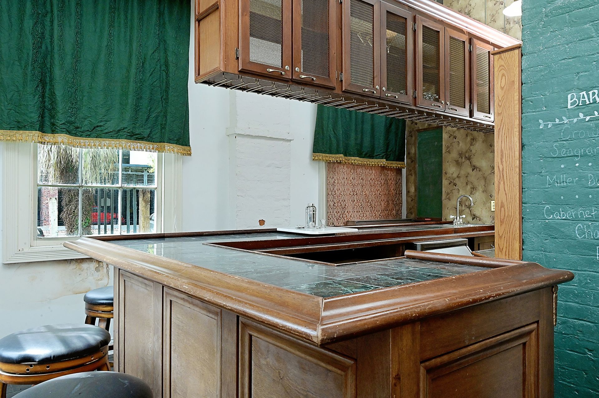 Wooden bar with stools, green curtains, and cabinets. A window and brick wall are visible.
