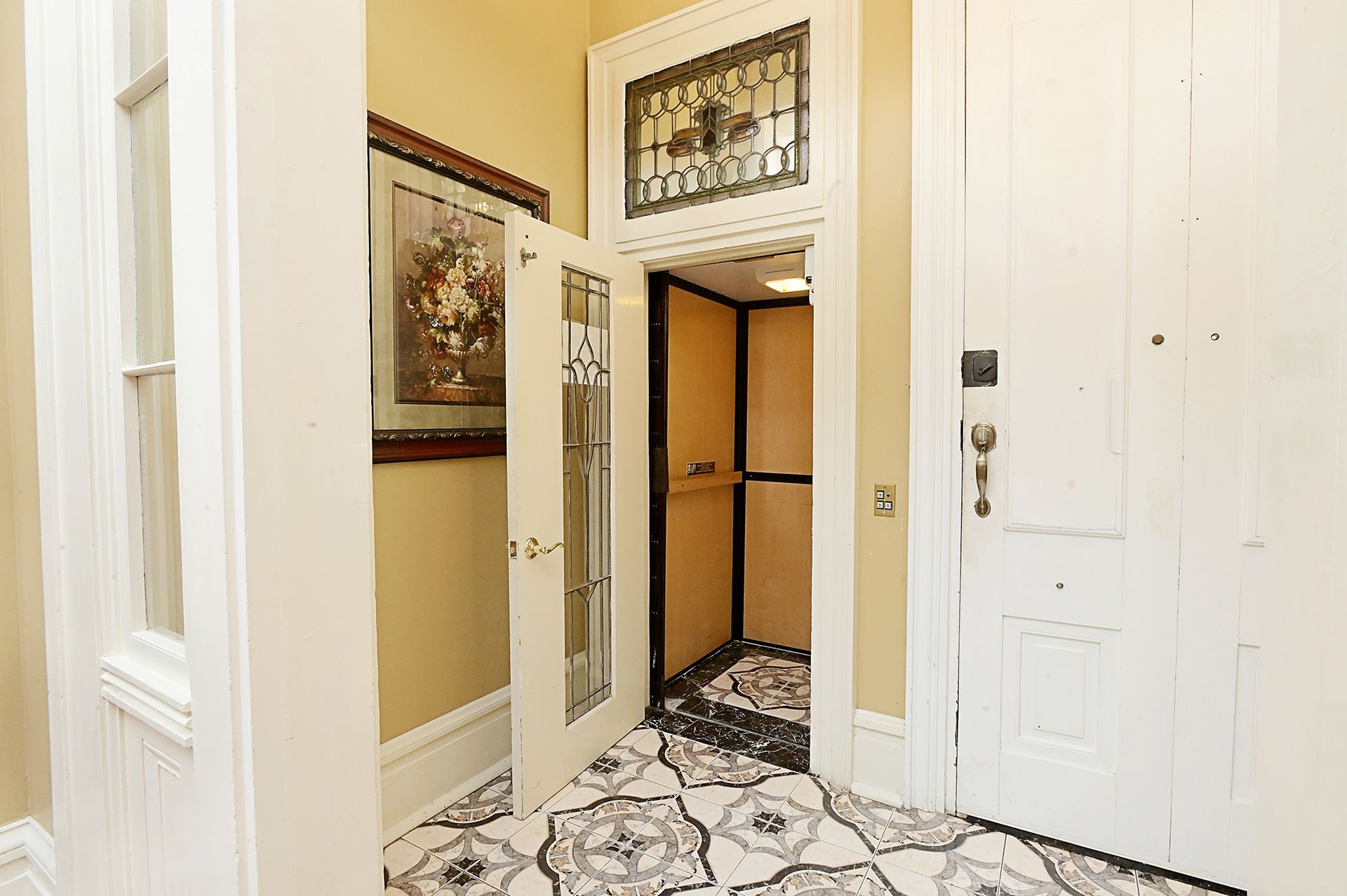 Hallway with elevator. Open elevator door reveals interior. Ornate tile floor. Decorative stained glass.