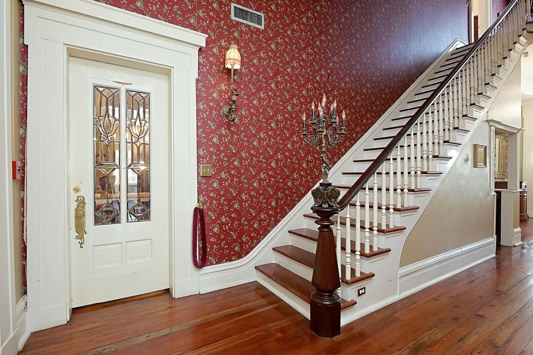 Interior entryway with staircase and ornate door. Red patterned wallpaper and wood floors.