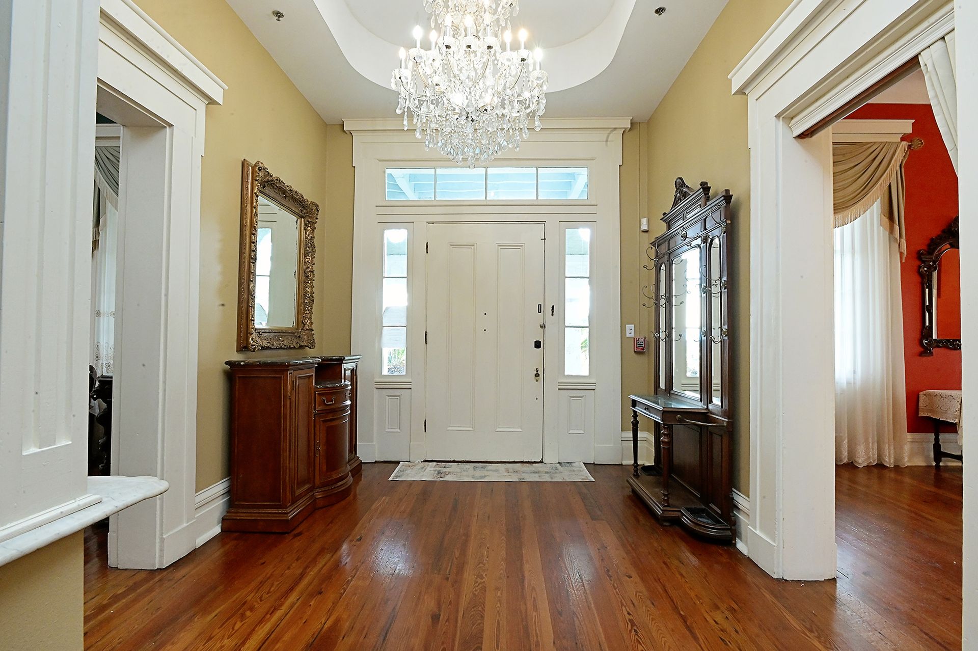 Elegant foyer with wood floors, chandelier, antique furniture, and white doorway.
