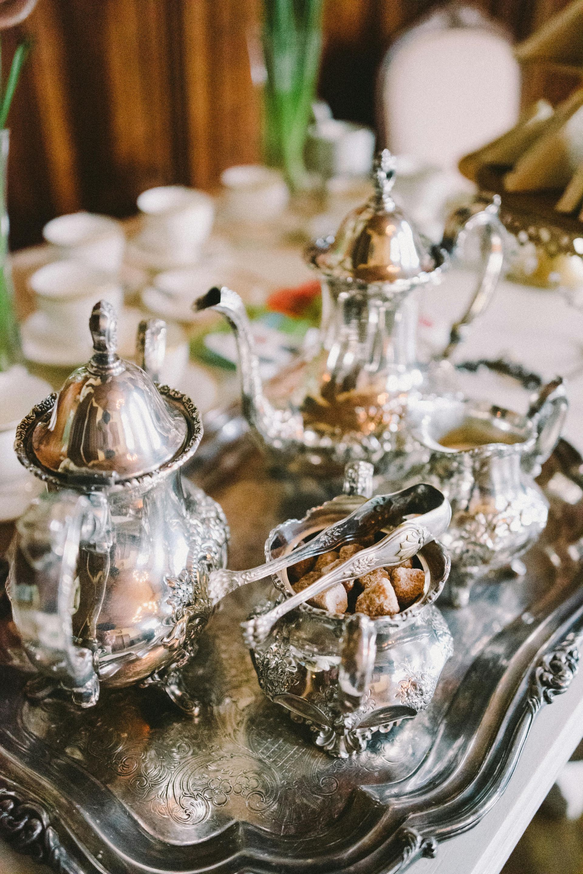 Silver tea set on a tray with sugar cubes. White teacups and flowers in background.