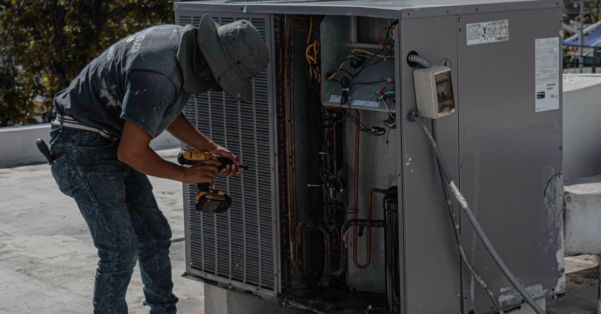 Un técnico repara una unidad exterior de climatización con herramientas, agachado junto a la maquinaria expuesta.