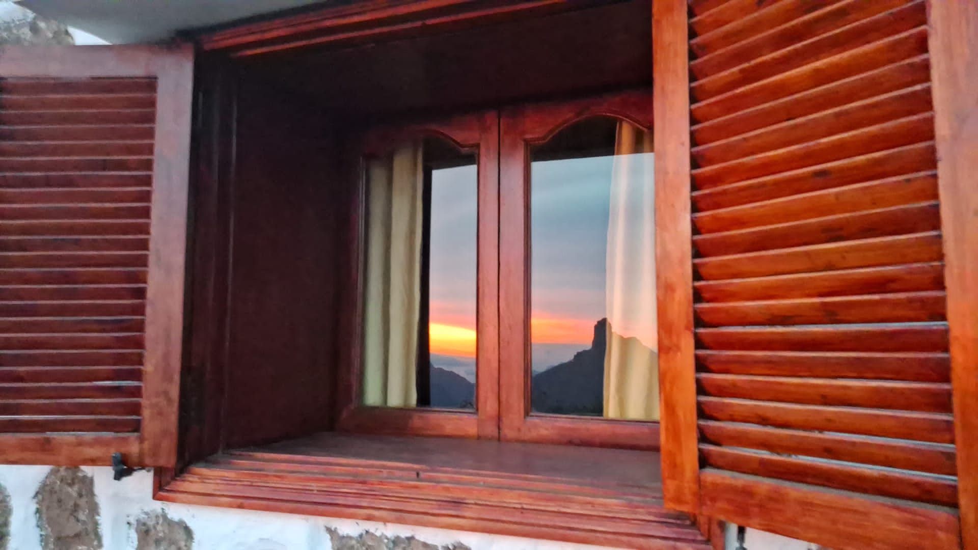 A window with wooden shutters is open to a view of the sunset.