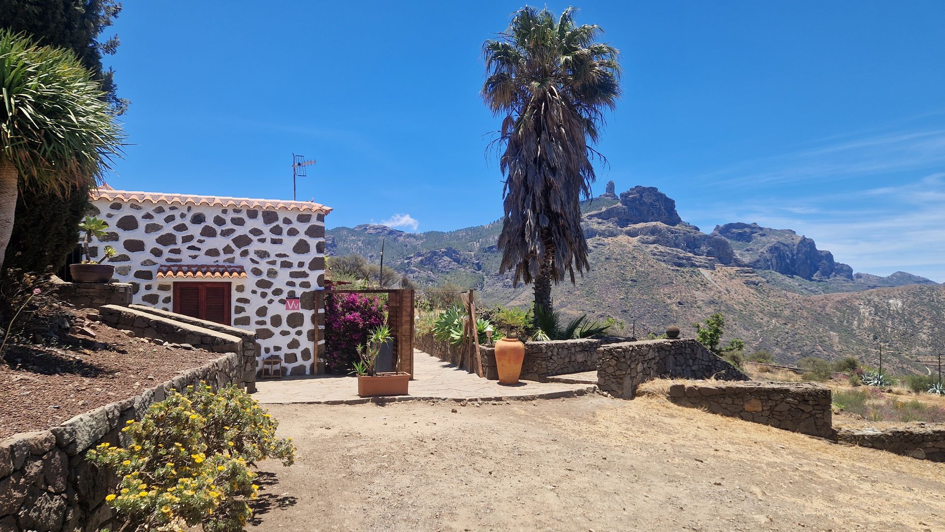 A stone house with a palm tree in front of it and mountains in the background.