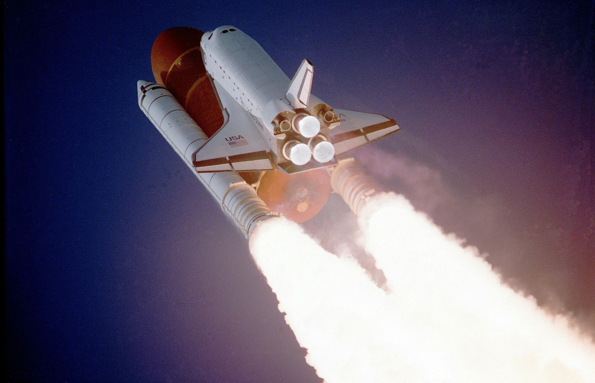 Space shuttle ascending with flames and exhaust, set against a dark blue sky.