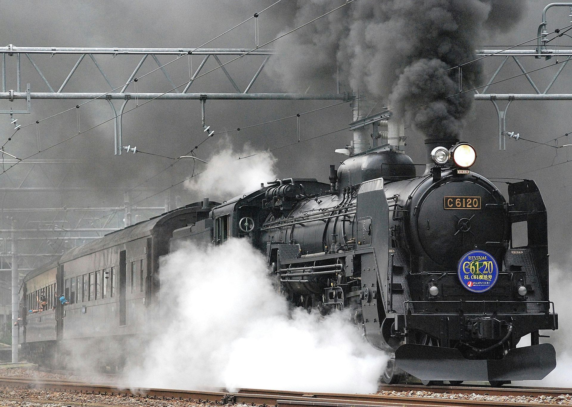 Steam train billowing black smoke, traveling along tracks, with passenger cars attached.