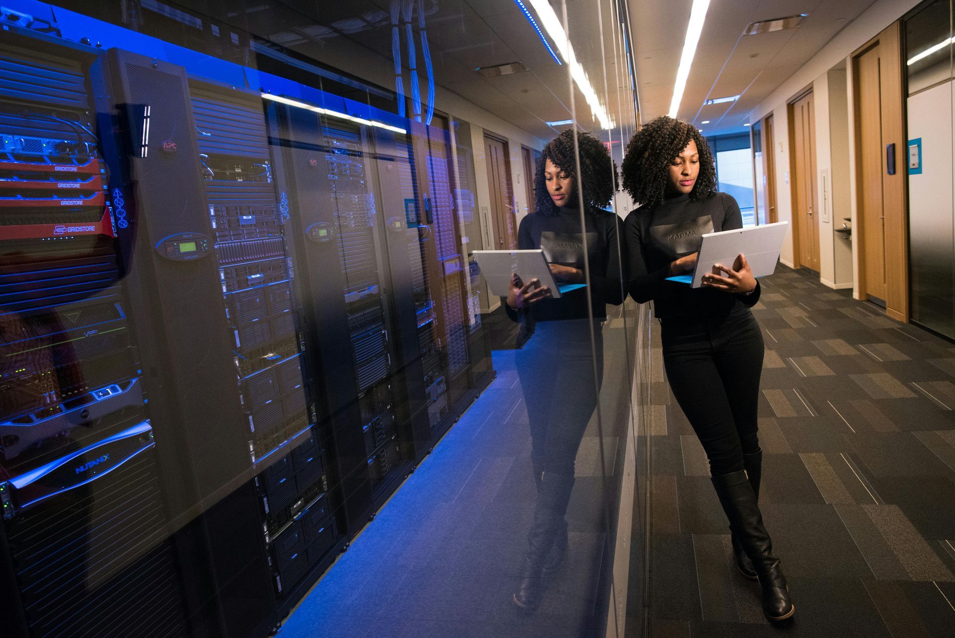 Woman in black attire standing near server racks, using a laptop in a modern data center corridor.