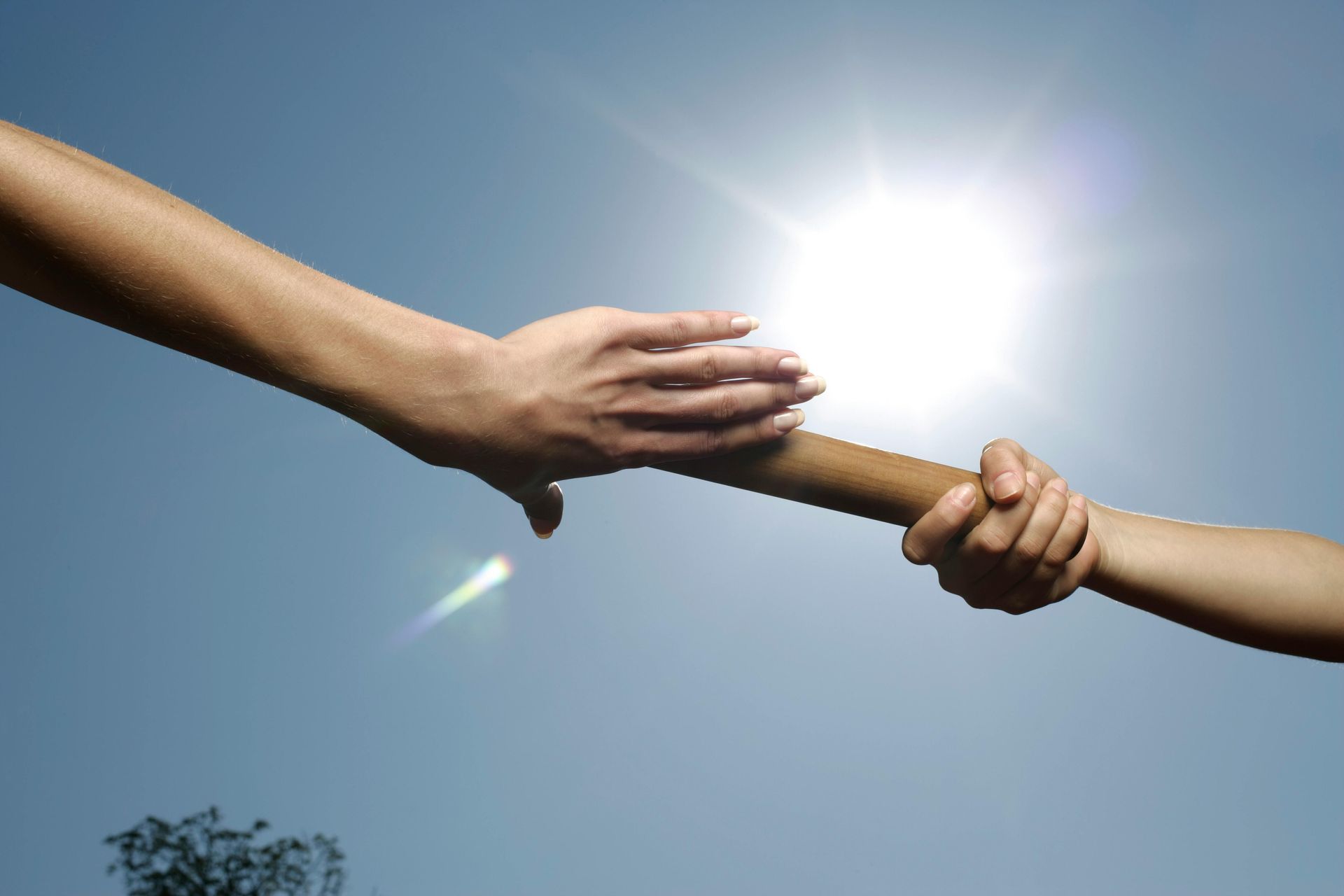 Hands passing a baton against a bright sunlit sky, symbolizing teamwork and relay race.