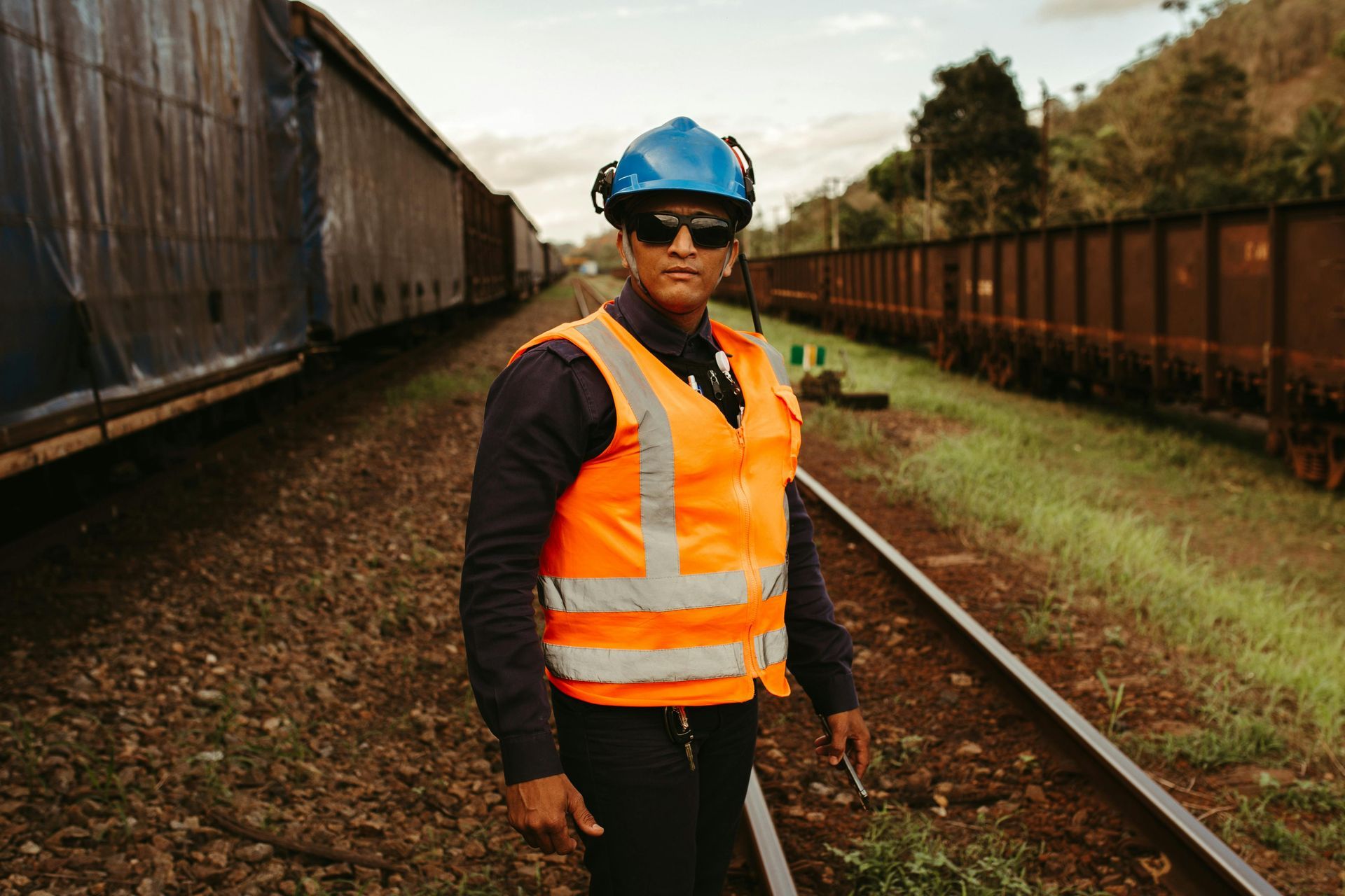 Man in safety gear stands on train tracks, between train cars, wearing sunglasses and a blue hard hat.