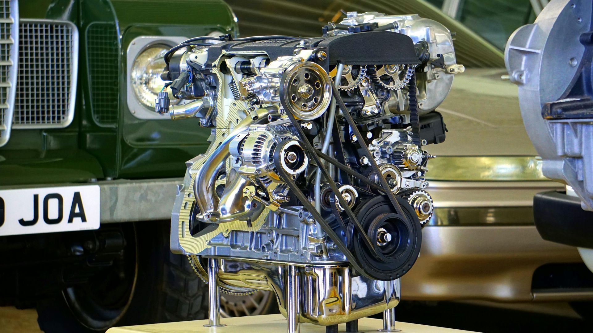 Chrome-plated engine displayed in front of vehicles, showcasing its internal mechanisms, belts, and gears.