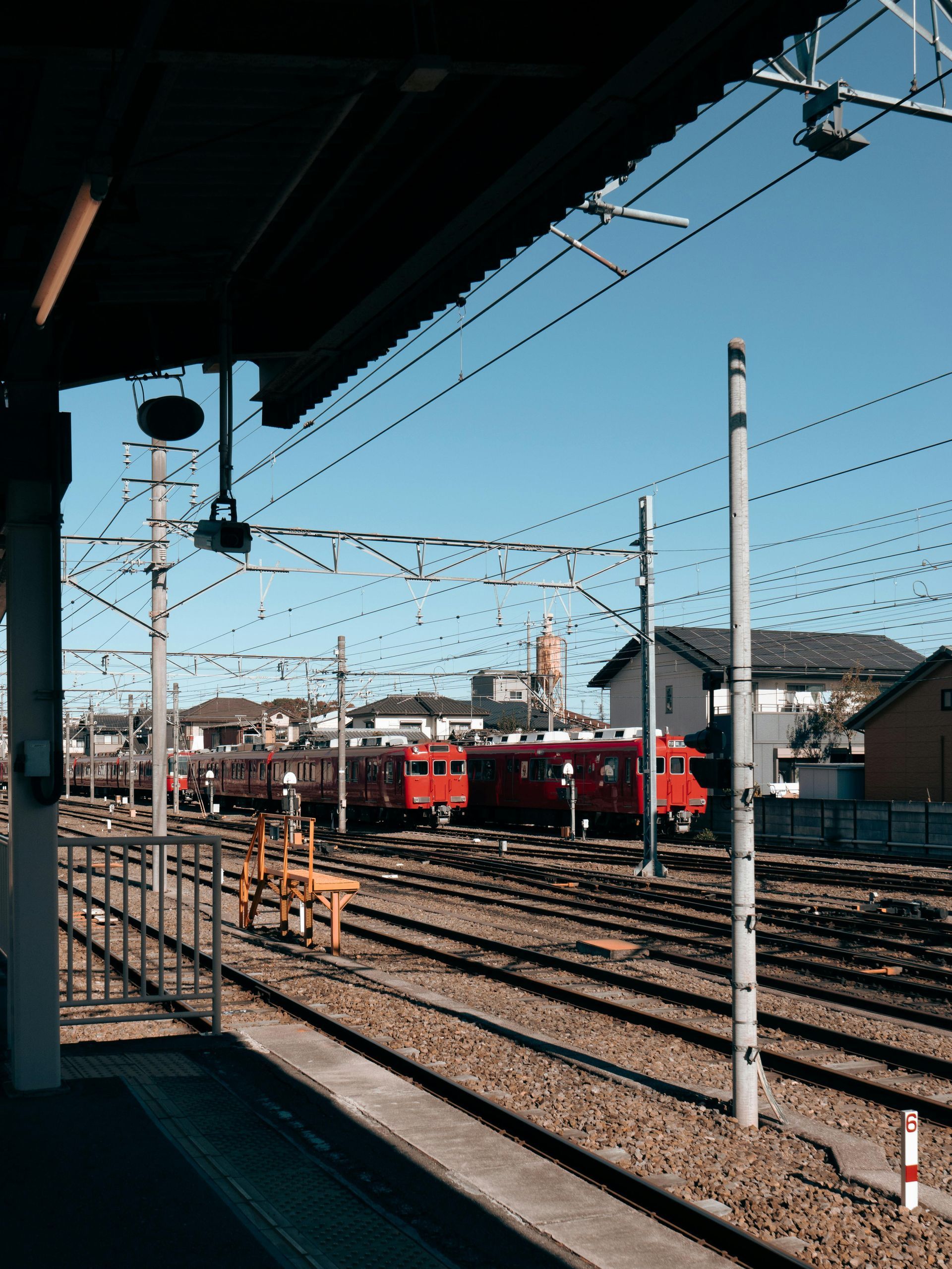 Red train on tracks at a sunny railway station, wires overhead, houses in background.