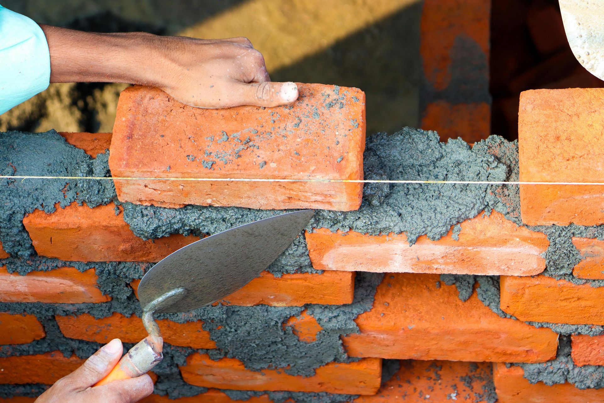 A hand laying a red brick on a wall with mortar, using a trowel; daylight.