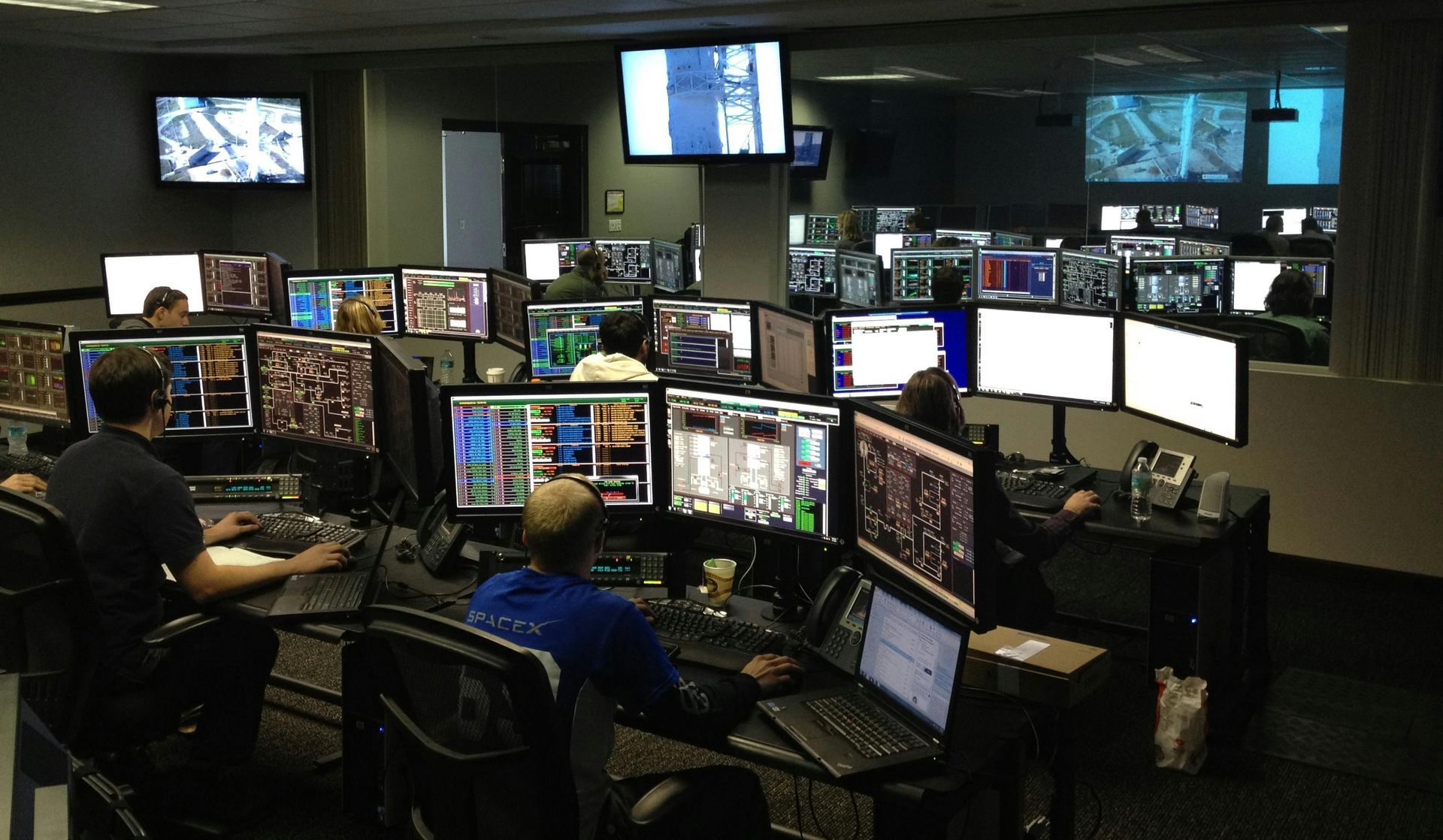 Control room with multiple monitors displaying data; people operate at consoles.