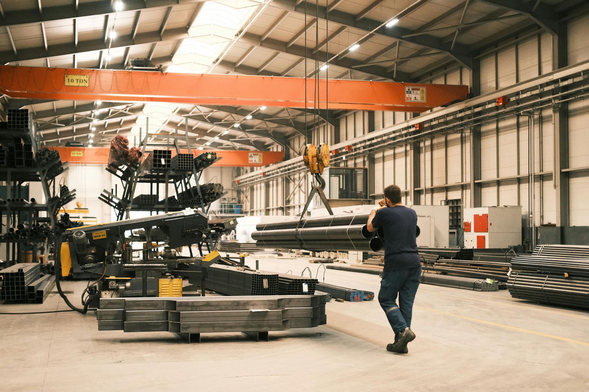 Man in a factory walks towards overhead crane lifting metal pipes. Industrial setting with equipment.